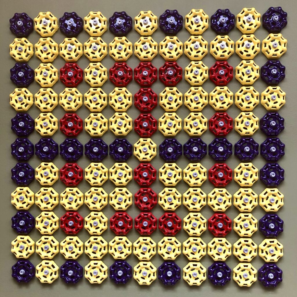 A square grid arrangement of octagonal faucet handles, creating an 11x11 pattern. The handles are in three colors: yellow (predominant), purple (along the edges and in specific patterns), and red (forming a cross or plus sign in the center). Each handle has a spoked wheel design and a central metal bolt. The handles appear to be made of plastic or painted metal and are photographed against a neutral grey background. The arrangement creates a symmetrical, decorative pattern reminiscent of pixel art or a quilt design. by James K-M